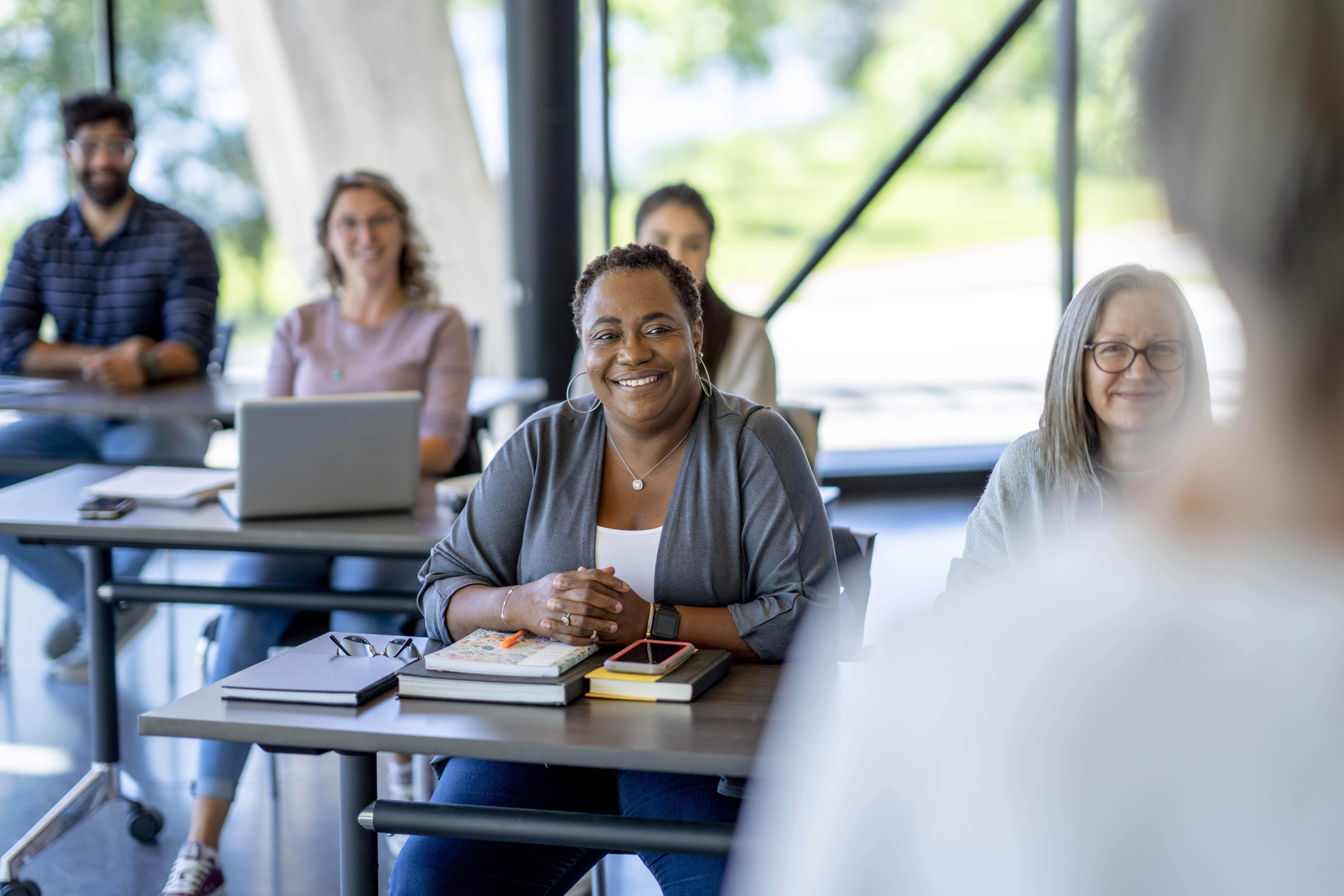 Women sitting at a table with books. People in background at tables with laptops and taking notes.