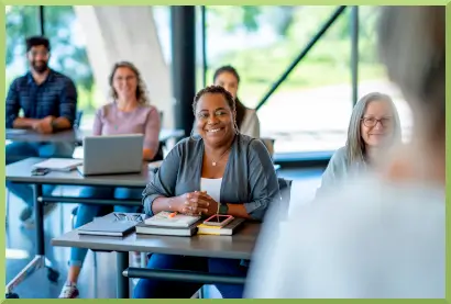 Women sitting at a table with books. People in background at tables with laptops and taking notes.
